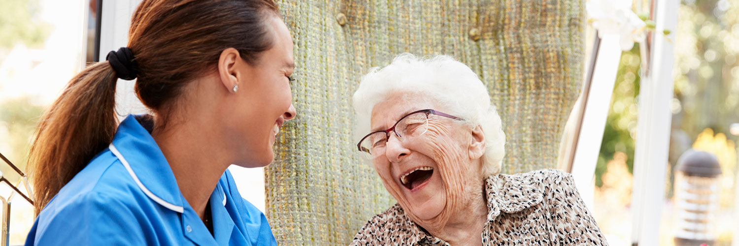 Senior Woman Sitting In Chair And Laughing With Nurse In Retirement Home